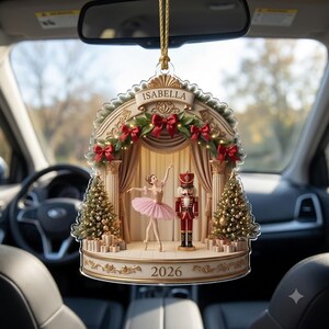 May include: A bell-shaped hanging ornament featuring a stage scene with a ballerina in a pink tutu and a nutcracker soldier. The archway is decorated with red bows and greenery, with the name ISABELLA and the year 2026 printed on the ornament.