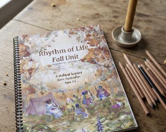 May include: A spiral-bound curriculum book titled Rhythm of Life Fall Unit, featuring a watercolor illustration of figures in a forest setting, accompanied by a collection of colored pencils and a single beeswax candle in a small ceramic holder.