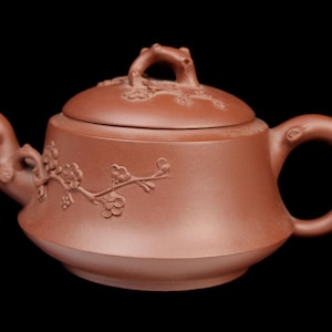 May include: A reddish-brown ceramic teapot with a lid, spout, and handle. The teapot features embossed floral designs and a curved handle. The spout is shaped like a bird's head. The lid has a frog-shaped handle.