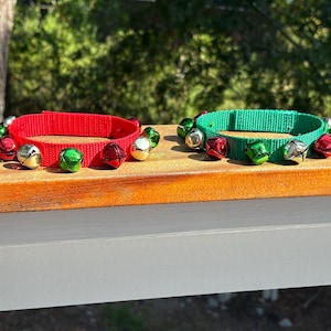 Może przedstawiać: Dwie świąteczne obroże dla zwierząt, jedna czerwona i jedna zielona, ​​każda ozdobiona serią małych, kolorowych dzwoneczków. Dzwonki są w odcieniach czerwieni, zieleni i srebra. Obroże są prezentowane na drewnianej powierzchni.