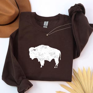 May include: A dark brown long-sleeve crewneck sweatshirt featuring a white, distressed-style graphic of a buffalo silhouette centered on the chest. The garment has a standard ribbed neckline and cuffs, with a Gildan brand label visible at the collar.