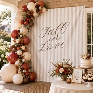 May include: A vertical balloon garland in shades of cream, tan, and deep burgundy, accented with dried foliage. A striped backdrop features the text Fall in Love. A floral centerpiece, a tiered cake, and a framed sign sit on a round table.