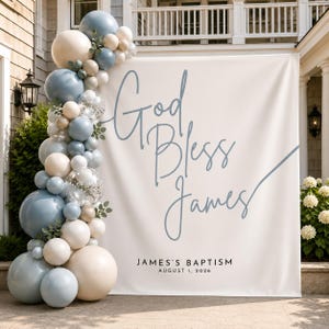 Pode incluir: Um grande pano de fundo branco com o texto em azul God Bless James e o texto em preto James's Baptism August 1, 2026. Uma guirlanda vertical de balões em tons de azul poeirento, creme e balões transparentes com confete está fixada à esquerda.