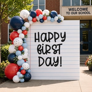 Könnte beinhalten: Ein großes rechteckiges Schild, das wie ein liniertes Blatt Notizpapier gestaltet ist und den schwarzen Text Happy First Day! trägt. Ein Ballonbogen in den Farben Marineblau, Rot, Weiß und Hellblau mit schwarzen Punkten rahmt das Schild ein.