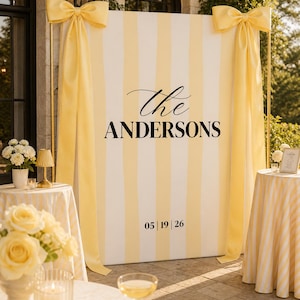 May include: A large rectangular event backdrop featuring vertical pale yellow and white stripes. The center displays the text "the ANDERSONS" in black, with the date "05 | 19 | 26" below. Two large yellow fabric bows are attached to the top corners.