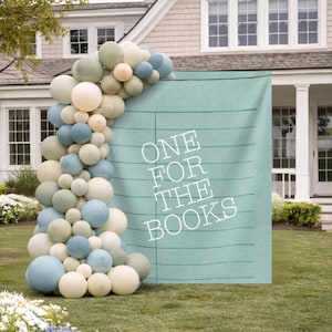 May include: A teal backdrop with the text "ONE FOR THE BOOKS" in white, decorated with a balloon arch in shades of blue, cream, and green. The scene is set outdoors in front of a house.