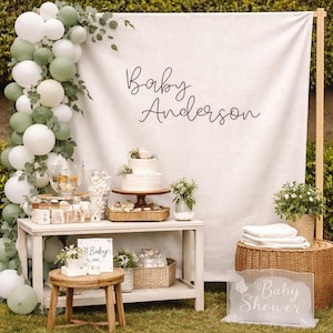 Op de afbeelding: Een baby shower achtergrond met een wit stoffen doek met de tekst Baby Anderson, versierd met een ballonnenboog in wit en saliegroen. Een houten tafel bevat een taart, witte blokken met de tekst BABY en een bordje met de tekst Oh Baby.