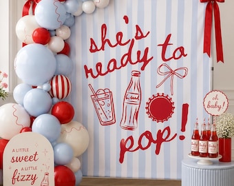 May include: A baby shower backdrop featuring light blue and white vertical stripes with the red text 'she's ready to pop!' and illustrations of a soda bottle, glass, and cap. A balloon arch in red, white, and light blue frames the left side.