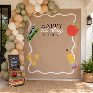 Peut inclure: Une toile de fond beige avec le texte HAPPY 1st day OF SCHOOL, ornée d'illustrations d'une pomme rouge, d'un crayon jaune et de deux craies. Une arche de ballons dans des tons beige, blanc et vert sauge encadre la scène avec un petit tableau.