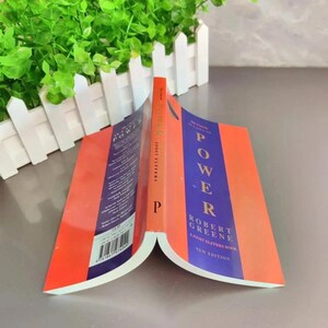 May include: An open book titled "The 48 Laws of Power" by Robert Greene. The book has an orange cover with the word "POWER" in large, purple letters. The spine is also orange with the letter "P".