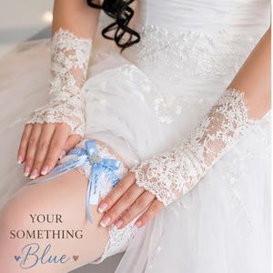 Op de afbeelding: Een bruidsbeen met een lichtblauwe kousenband en witte kanten handschoenen. De kousenband heeft een blauwe strik met een sprankelend middelpunt en een gepersonaliseerd lint. De tekst "YOUR SOMETHING Blue" is zichtbaar.