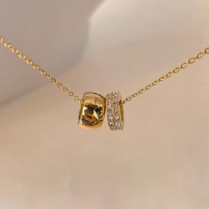 May include: A gold-colored necklace featuring two connected ring pendants. One ring is solid gold, while the other is adorned with small, sparkling crystals. The necklace hangs on a delicate gold chain, creating a simple yet elegant design.