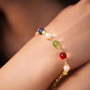 Könnte beinhalten: Ein goldenes Armband mit einer Reihe farbiger Perlen, darunter blaue, weiße, grüne und rote. Das Armband wird an einem Handgelenk getragen, wodurch das Design und die Farbvariationen hervorgehoben werden. Die Perlen haben einen Durchmesser von etwa 0,8 cm.