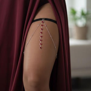 May include: A silver leg garter with a black elastic band and a chain design. The chain is decorated with small, red heart-shaped charms. The garter is worn on the upper thigh, adding a touch of elegance. The garter is a stylish accessory.