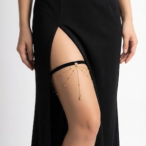 May include: A gold-coloured leg garter with a black velvet band. The garter features delicate chains and small beads, with a dangling initial charm. The garter is worn on the thigh, and is paired with a black dress.