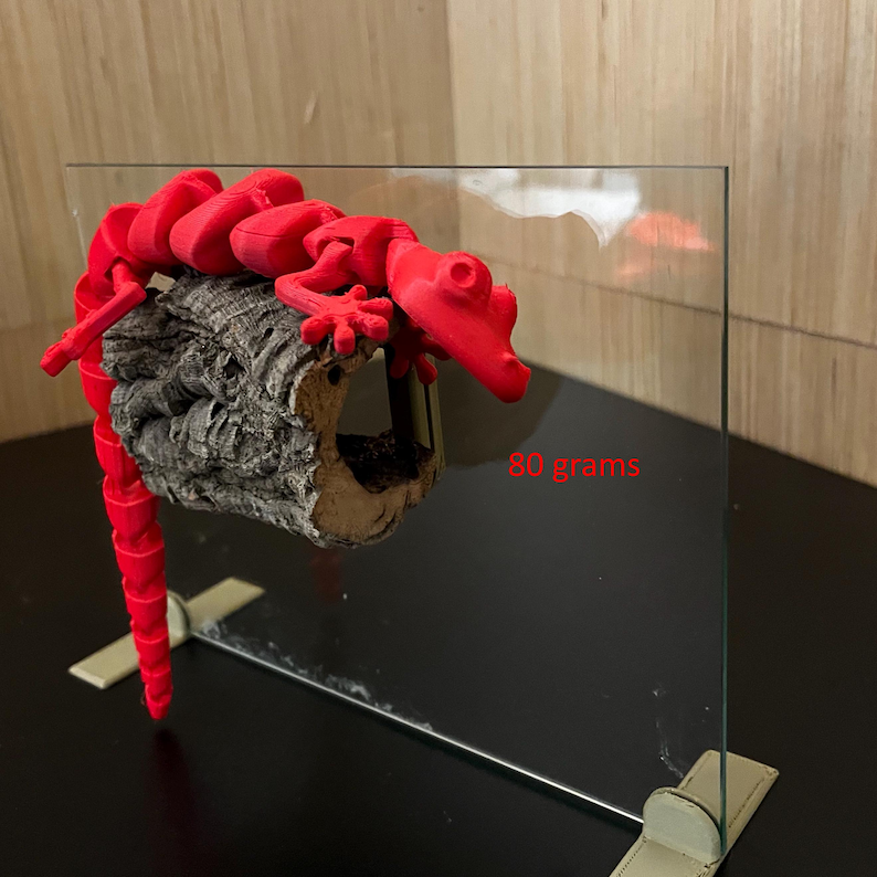 K&ouml;nnte beinhalten: Eine leuchtend rote, bewegliche Eidechsenfigur, die auf einem St&uuml;ck strukturierter, grauer Rinde sitzt. Die Eidechse ist 3D-gedruckt und scheint sich auf der Rinde auszuruhen. Der Text "80 Gramm" ist im Bild zu sehen.