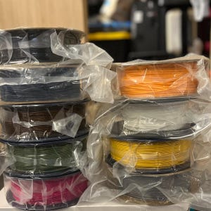 K&ouml;nnte beinhalten: Ein Stapel von 3D-Drucker-Filamentrollen in verschiedenen Farben, darunter Schwarz, Orange, Wei&szlig;, Gelb, Gr&uuml;n und Pink. Jede Rolle ist in klarem Kunststoff verpackt. Die Rollen sind &uuml;bereinander gestapelt.