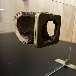 K&ouml;nnte beinhalten: Eine naturalistische Terrariendekoration mit einem ausgeh&ouml;hlten Baumstammabschnitt und einem olivgr&uuml;nen Rahmen. Der Baumstamm hat eine raue, strukturierte Oberfl&auml;che mit verschiedenen Braunt&ouml;nen. Der Rahmen hat einen dekorativen, gen&auml;hten Rand und eine quadratische &Ouml;ffnung.