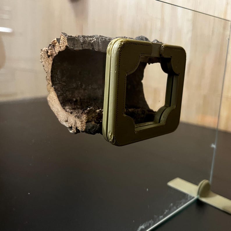 K&ouml;nnte beinhalten: Eine naturalistische Terrariendekoration mit einem ausgeh&ouml;hlten Baumstammabschnitt und einem olivgr&uuml;nen Rahmen. Der Baumstamm hat eine raue, strukturierte Oberfl&auml;che mit verschiedenen Braunt&ouml;nen. Der Rahmen hat einen dekorativen, gen&auml;hten Rand und eine quadratische &Ouml;ffnung.