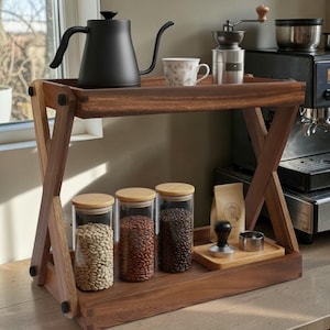 Puede incluir: Una estación de café de madera con una tetera de cuello de cisne negra, una taza de té floral y un molinillo de café manual en el estante superior. El estante inferior contiene frascos de vidrio con granos de café.