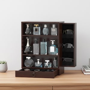 May include: A dark brown wooden perfume and jewellery organiser with multiple shelves and a drawer. The shelves display various perfume bottles of different shapes and sizes. The right side has a door with slots for sunglasses. The drawer is open, revealing small compartments.