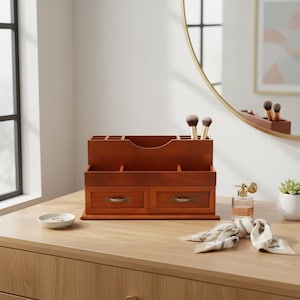 May include: A wooden makeup organizer with two drawers and multiple compartments. The organizer is a warm brown color and has brass-colored drawer pulls. Makeup brushes are placed in the top section. The organizer sits on a wooden surface.