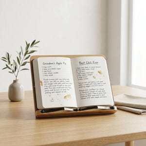 May include: A wooden cookbook stand holds an open notebook with handwritten recipes. The left page reads "Grandma's Apple Pie," and the right page says "Best Chili Ever." The stand is on a light-colored wooden table, with a small vase and a sprig of greenery.