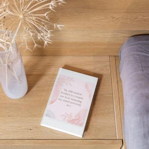 May include: A white affirmation booklet with pink and gray watercolor designs and text, resting on a wooden surface. A clear vase with dried flowers is to the left, and a bed with a gray comforter is on the right.