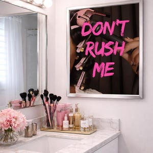 May include: A bathroom vanity with a mirror displaying the text "DON'T RUSH ME" in pink. The vanity features a marble countertop, a glass vase with pink flowers, makeup brushes, and various beauty products.