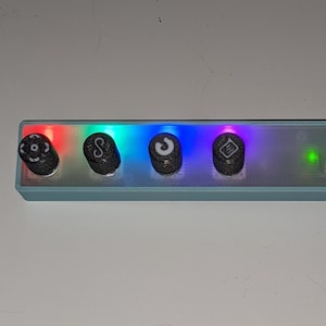 Op de afbeelding: Een rechthoekig, lichtblauw elektronisch apparaat met vier zwarte knoppen, elk met een uniek symbool. Het apparaat heeft een regenboogkleurige lichtweergave en een groen indicatielampje. Een zwarte kabel is aangesloten.