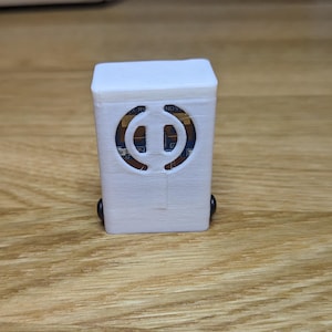 Può includere: Un piccolo oggetto rettangolare bianco con un ritaglio decorativo sul davanti, che rivela i componenti interni. L'oggetto ha piccole ruote nere sul fondo. L'articolo è appoggiato su una superficie di legno.