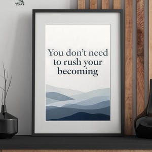 May include: Framed art print with a black border. The print features the text "You don't need to rush your becoming" in a dark blue font, above a landscape of blue and gray layered hills. The Fine Urban Aura logo is in the top left corner.