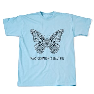 May include: A light blue t-shirt featuring a black butterfly design filled with floral patterns. The text "TRANSFORMATION IS BEAUTIFUL" is printed below the butterfly. The shirt is made of a soft material.