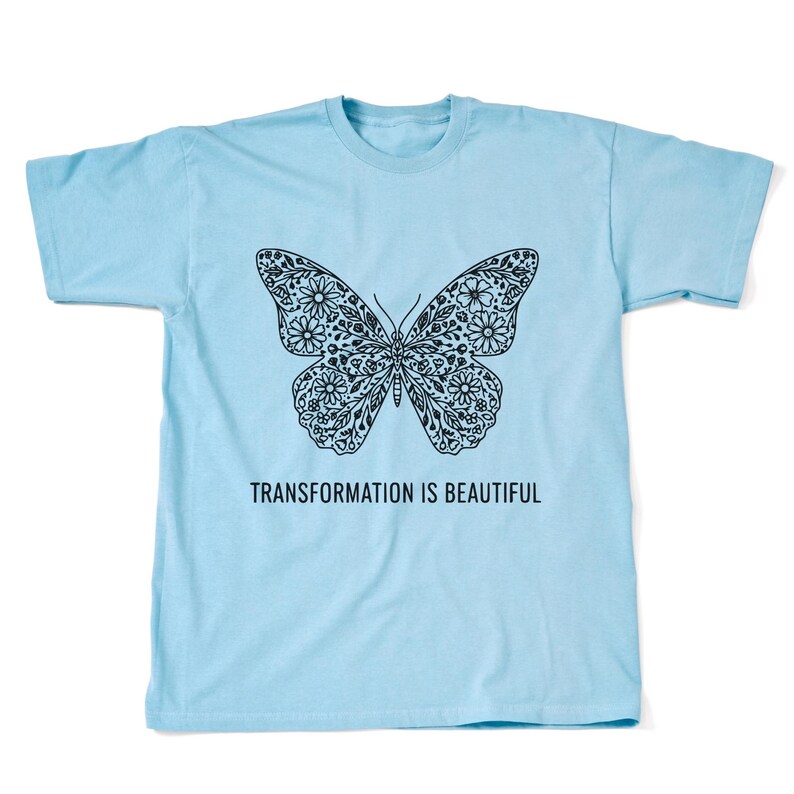 May include: A light blue t-shirt featuring a black butterfly design filled with floral patterns. The text "TRANSFORMATION IS BEAUTIFUL" is printed below the butterfly. The shirt is made of a soft material.