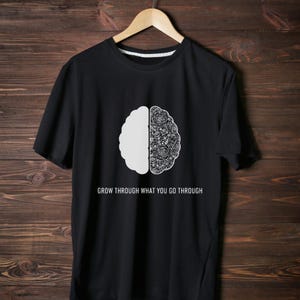 May include: Black t-shirt with a white brain graphic. One half of the brain is solid, the other half is a floral design. The text "GROW THROUGH WHAT YOU GO THROUGH" is printed below the graphic.