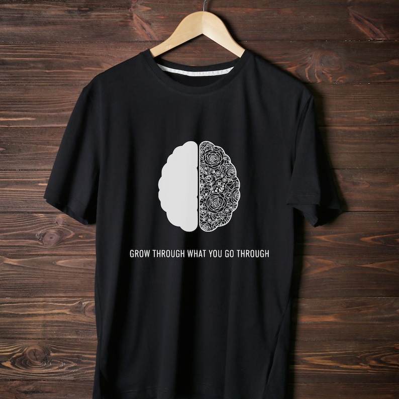 May include: Black t-shirt with a white brain graphic. One half of the brain is solid, the other half is a floral design. The text "GROW THROUGH WHAT YOU GO THROUGH" is printed below the graphic.