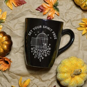 May include: Black ceramic mug with a curved handle, featuring the text "SET YOUR SPIRIT FREE" and a white illustration of a birdcage with floral accents. The mug is surrounded by fall-themed decorations, including pumpkins and leaves.