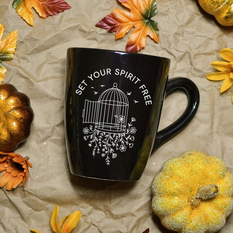 May include: Black ceramic mug with a curved handle, featuring the text "SET YOUR SPIRIT FREE" and a white illustration of a birdcage with floral accents. The mug is surrounded by fall-themed decorations, including pumpkins and leaves.