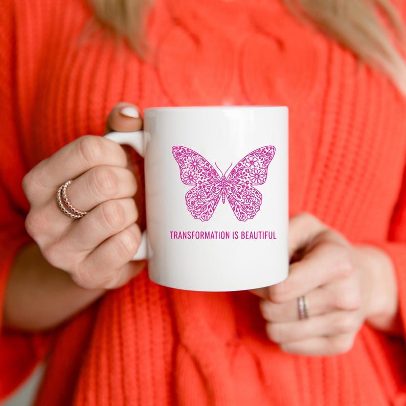 May include: White ceramic mug featuring a magenta butterfly design with floral patterns. The mug has the text "TRANSFORMATION IS BEAUTIFUL" printed below the butterfly. The mug is held by a person wearing a red sweater.