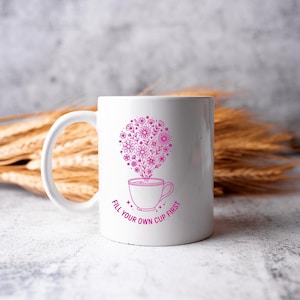 May include: White ceramic mug with a pink floral design and the text "FILL YOUR OWN CUP FIRST". The design features a cup with flowers rising from it. The mug has a curved handle.