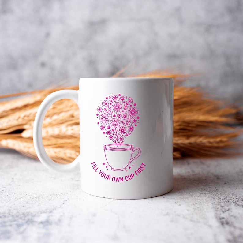 May include: White ceramic mug with a pink floral design and the text "FILL YOUR OWN CUP FIRST". The design features a cup with flowers rising from it. The mug has a curved handle.