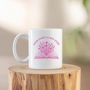 May include: White ceramic mug with a pink floral design and the text "YOUR STORY IS NOT OVER". The design features an open book with flowers and foliage. The mug is on a wooden surface.