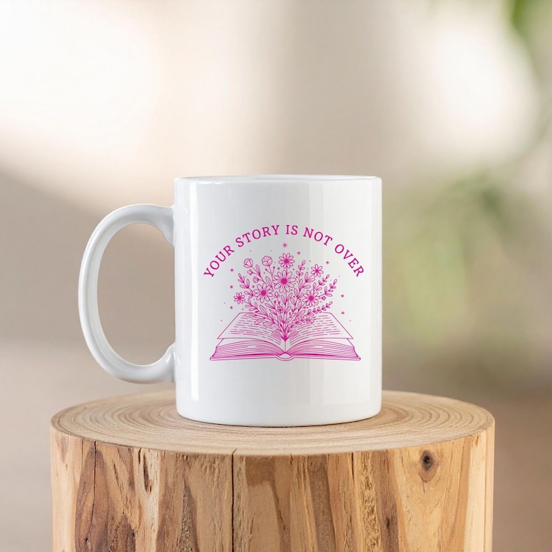 May include: White ceramic mug with a pink floral design and the text "YOUR STORY IS NOT OVER". The design features an open book with flowers and foliage. The mug is on a wooden surface.