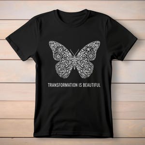 May include: Black t-shirt featuring a detailed white butterfly design. The butterfly's wings are filled with floral patterns. The text "TRANSFORMATION IS BEAUTIFUL" is printed below the butterfly.