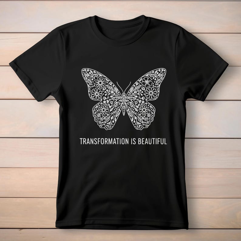 May include: Black t-shirt featuring a detailed white butterfly design. The butterfly's wings are filled with floral patterns. The text "TRANSFORMATION IS BEAUTIFUL" is printed below the butterfly.