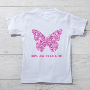 May include: White t-shirt featuring a detailed magenta butterfly design on the back. The butterfly is composed of floral patterns. Below the butterfly, the text reads "TRANSFORMATION IS BEAUTIFUL".