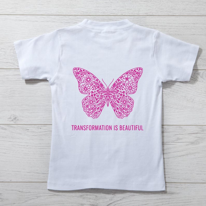 May include: White t-shirt featuring a detailed magenta butterfly design on the back. The butterfly is composed of floral patterns. Below the butterfly, the text reads "TRANSFORMATION IS BEAUTIFUL".