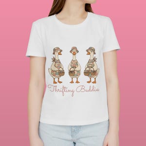 Può includere: T-shirt bianca con un'illustrazione stravagante di tre oche con cappelli e grembiuli, ognuna con un cestino. Il testo "Thrifting Buddies" è scritto in corsivo sotto.