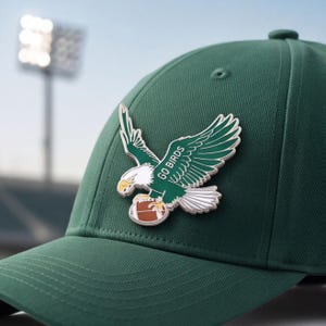Könnte beinhalten: Eine dunkelgrüne Baseballkappe mit einer metallischen Emaille-Anstecknadel auf der Vorderseite. Die Anstecknadel zeigt einen weißen und grünen Adler im Flug, der einen braunen Football hält, mit dem Text GO BIRDS auf dem Flügel.