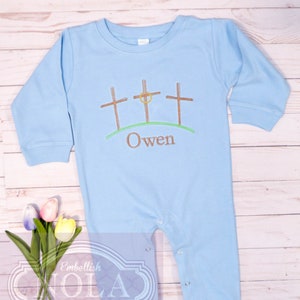 May include: A light blue baby onesie with embroidered brown crosses and a green hill. The name "Owen" is embroidered below the crosses.